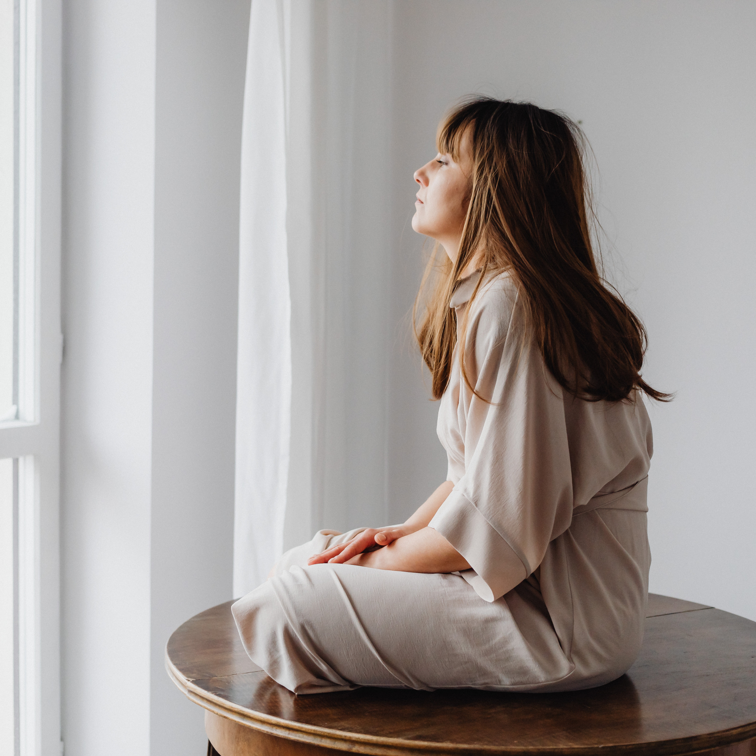 A woman sitting on a wooden table by a window with light curtains, in soft natural light, slightly withdrawn, calm but clearly not okay
