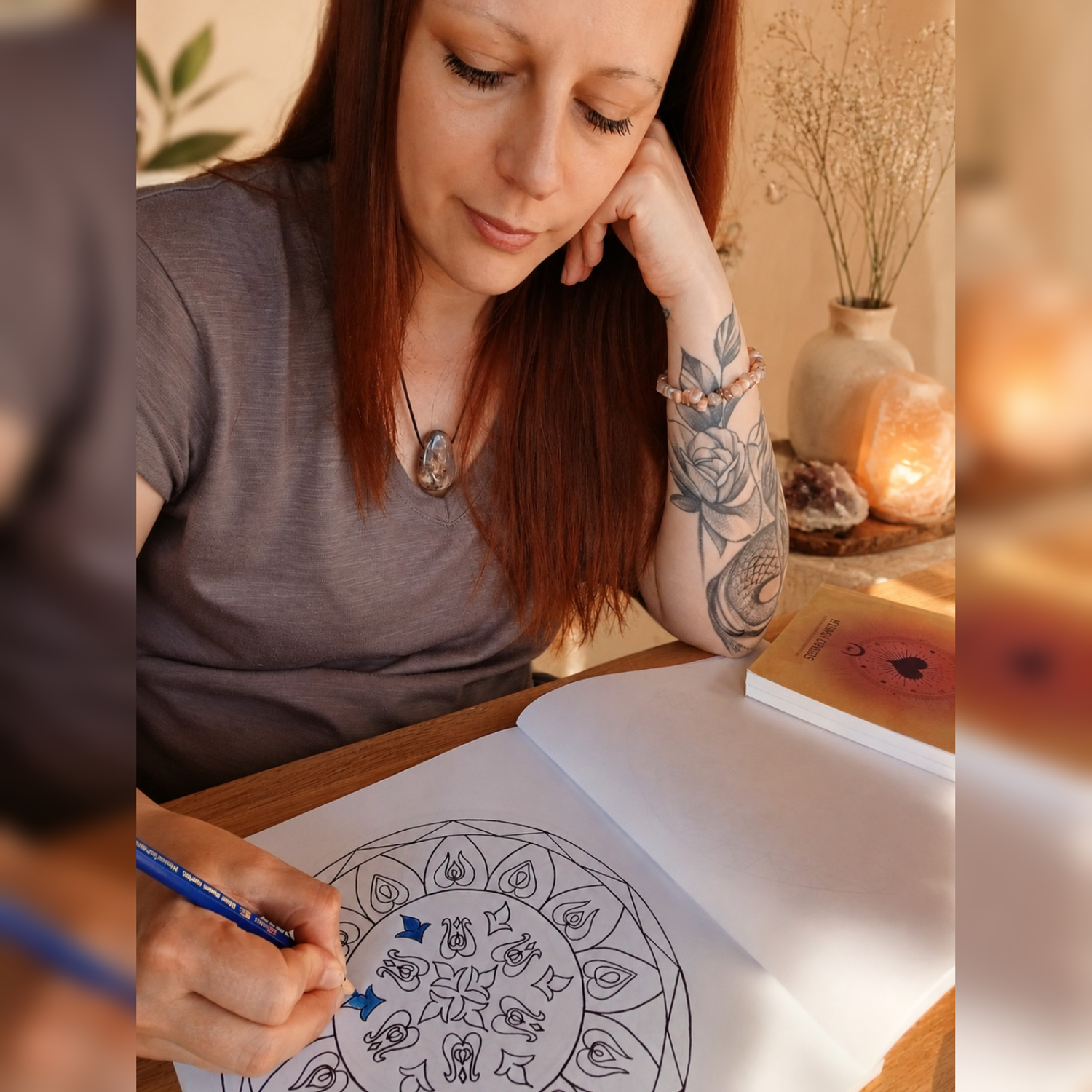 A woman with tattoos and red hair sits at a wooden table, coloring a hand-drawn mandala from the Coloring Wheels coloring book with a blue pencil. She is wearing a gray t-shirt and has a warm, cozy background with a glowing lamp and decorative plants.